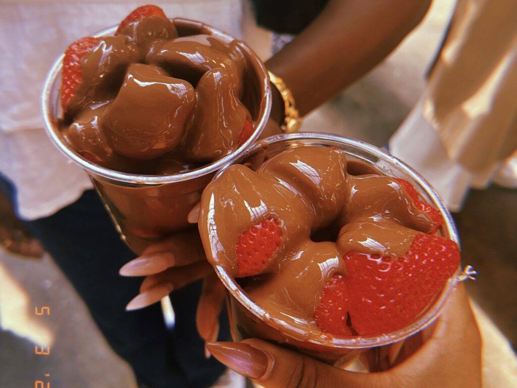 The hands of two Black girls holding  cups of chocolate covered strawberries.