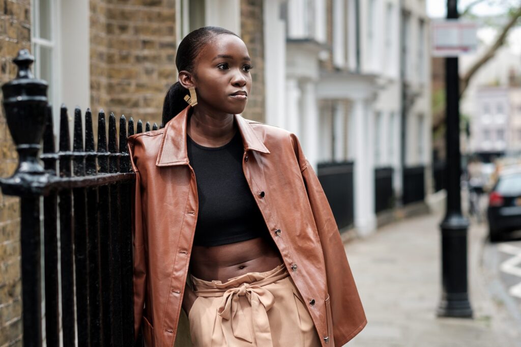 Stylish woman in brown leather jacket leaning on iron gate in the street