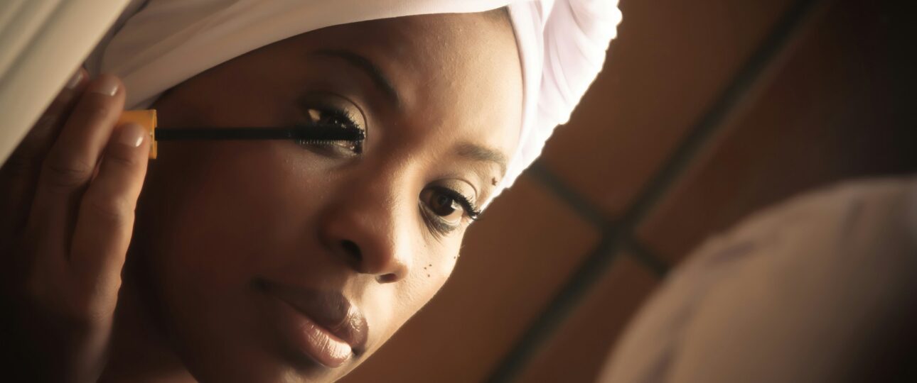 A dark skinned Black woman looking at herself in a mirror while putting on mascara. She's wearing a white towel wrapped around her head and a white robe.