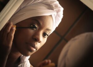 A dark skinned Black woman looking at herself in a mirror while putting on mascara. She's wearing a white towel wrapped around her head and a white robe.